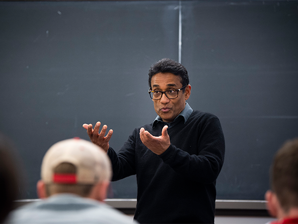 An instructor stands in front of a classroom, back against a blackboard, and speaks.